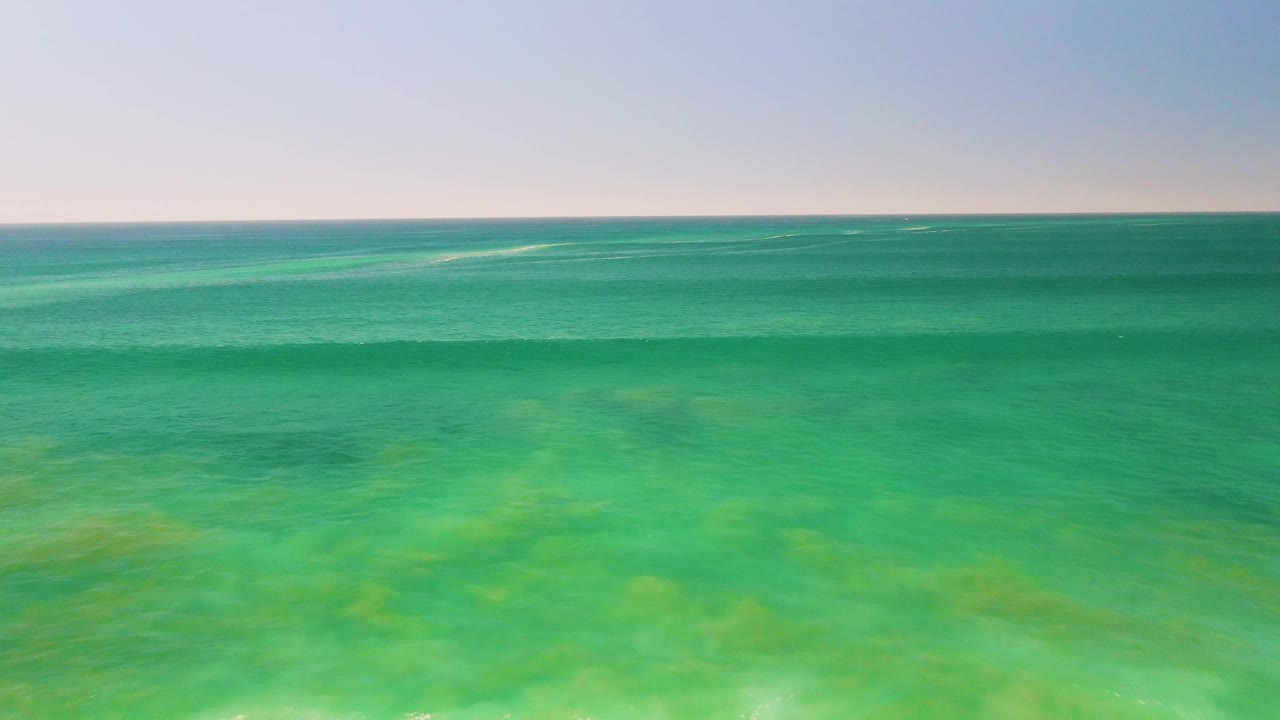 Wide aerial view of shallow turquoise water in San Jose del Cabo with green seabed tones, a distant level horizon, pale sky, and no visible people.