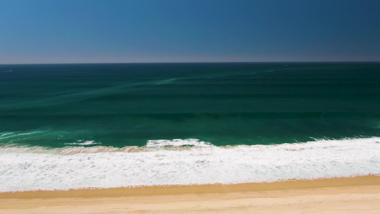 Aerial frame over San Jose del Cabo showing turquoise ocean, white surf, tan sand, a level horizon, and no visible people.