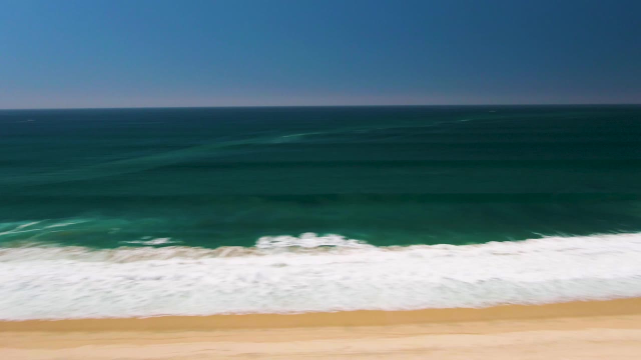 Aerial San Jose del Cabo shoreline view with darker offshore water bands, white breaking waves, pale sand, and no visible people.