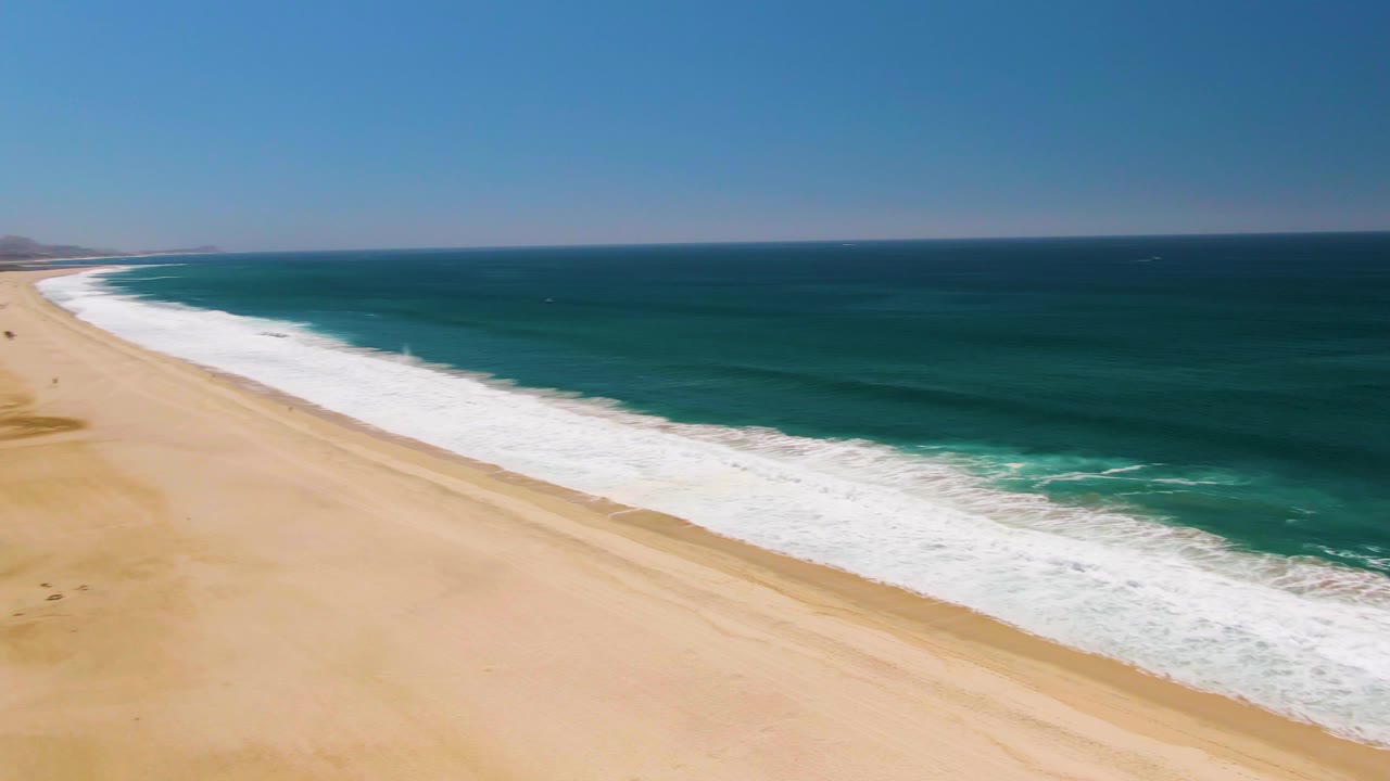 High aerial view in San Jose del Cabo looking down a long sandy beach with turquoise water on the right, distant coastline, and no visible people.