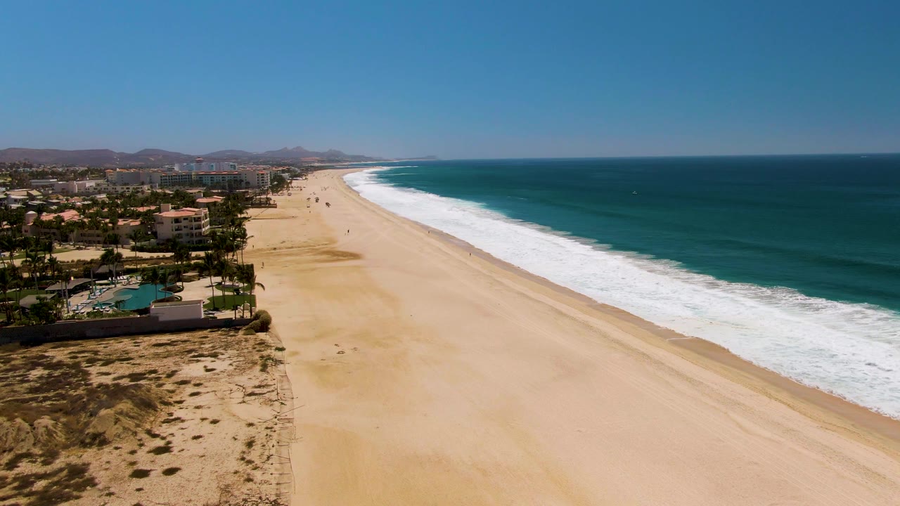 Aerial coastline with buildings, dunes, beach, and ocean surf.