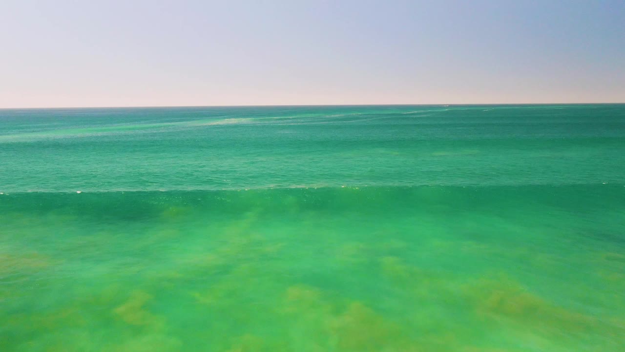 Wide aerial view of turquoise shallow ocean water in San Jose del Cabo, Mexico, with a low wave beginning to break near the foreground and no visible people.