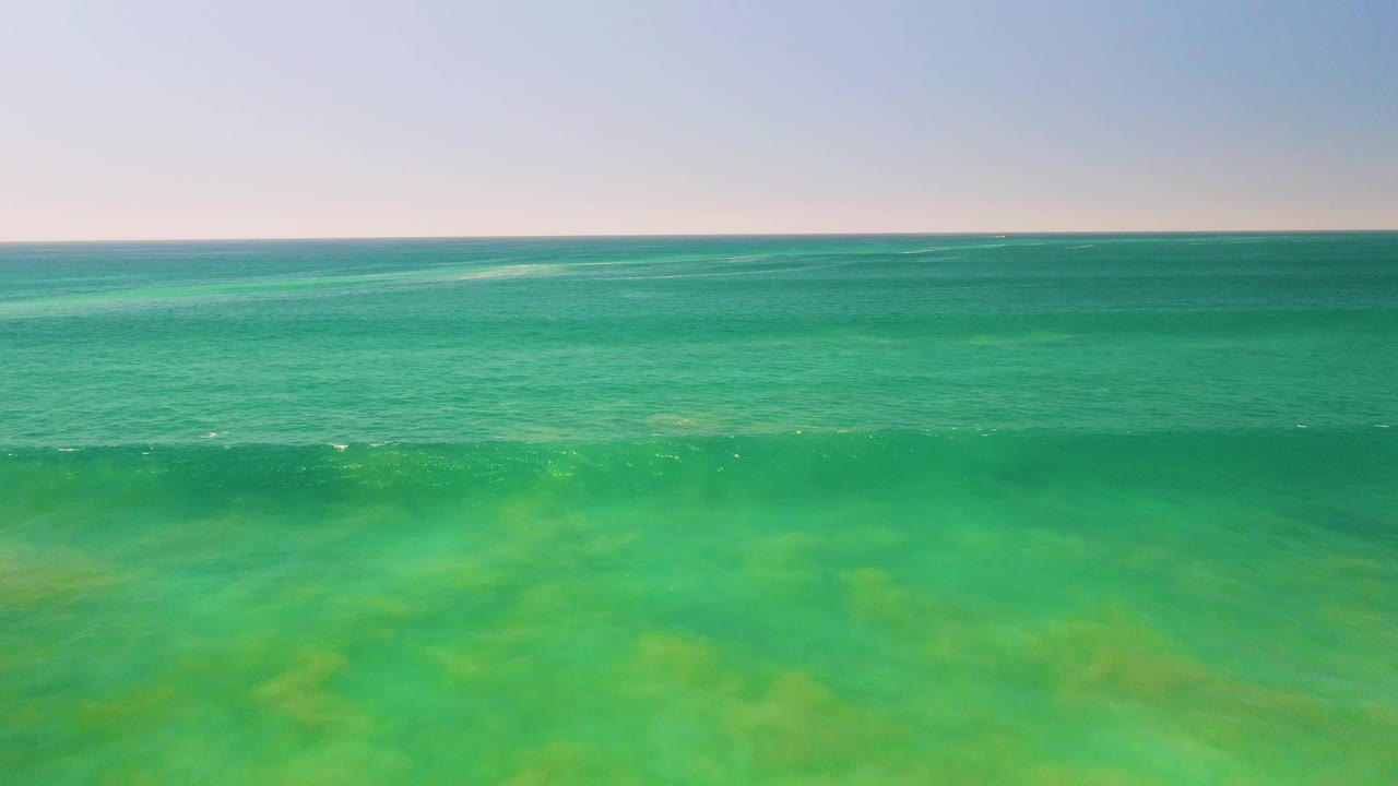 Aerial frame of clear green-blue shallows in San Jose del Cabo, Mexico, with faint white foam spreading across the lower water and no visible people.