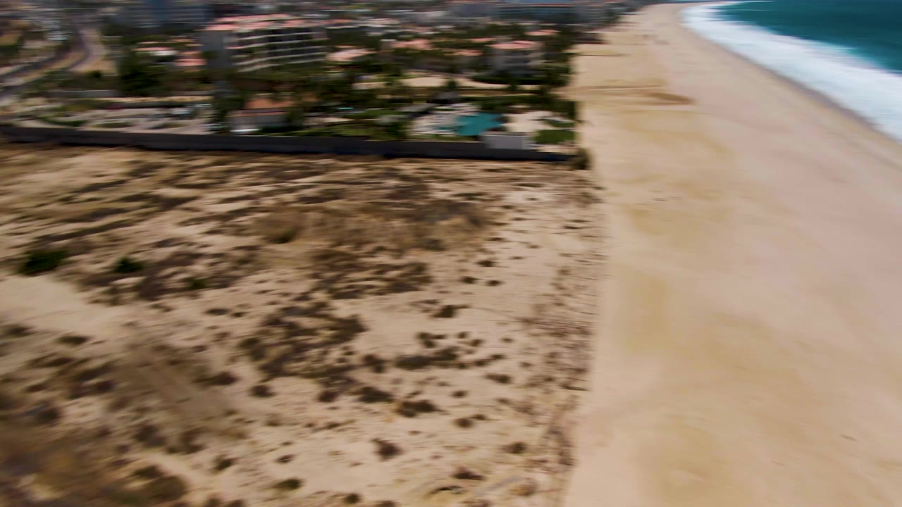 Aerial view in San Jose del Cabo showing a sandy beach and blue surf beside dry dunes and beachfront buildings, with motion blur and no visible people.