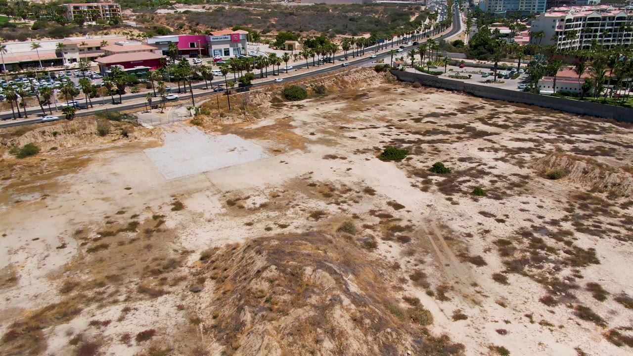 High aerial frame in San Jose del Cabo looking over eroded sandy dunes, a road lined with palms, parked or moving vehicles, and low buildings, with no visible people.