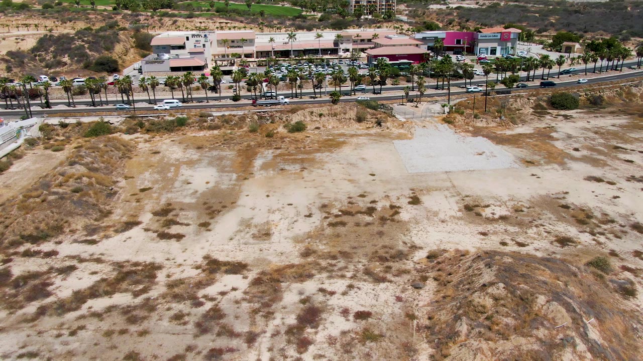 Wide drone view in San Jose del Cabo showing dry sandy terrain with scrub patches in the foreground, roads and palms through the middle, and beachfront buildings beyond, with no visible people.