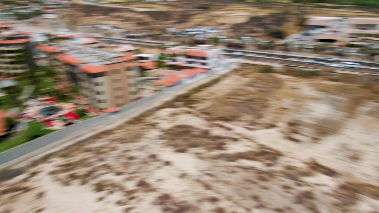 Blurred aerial pass over dunes, road, palm trees, and beachfront structures.
