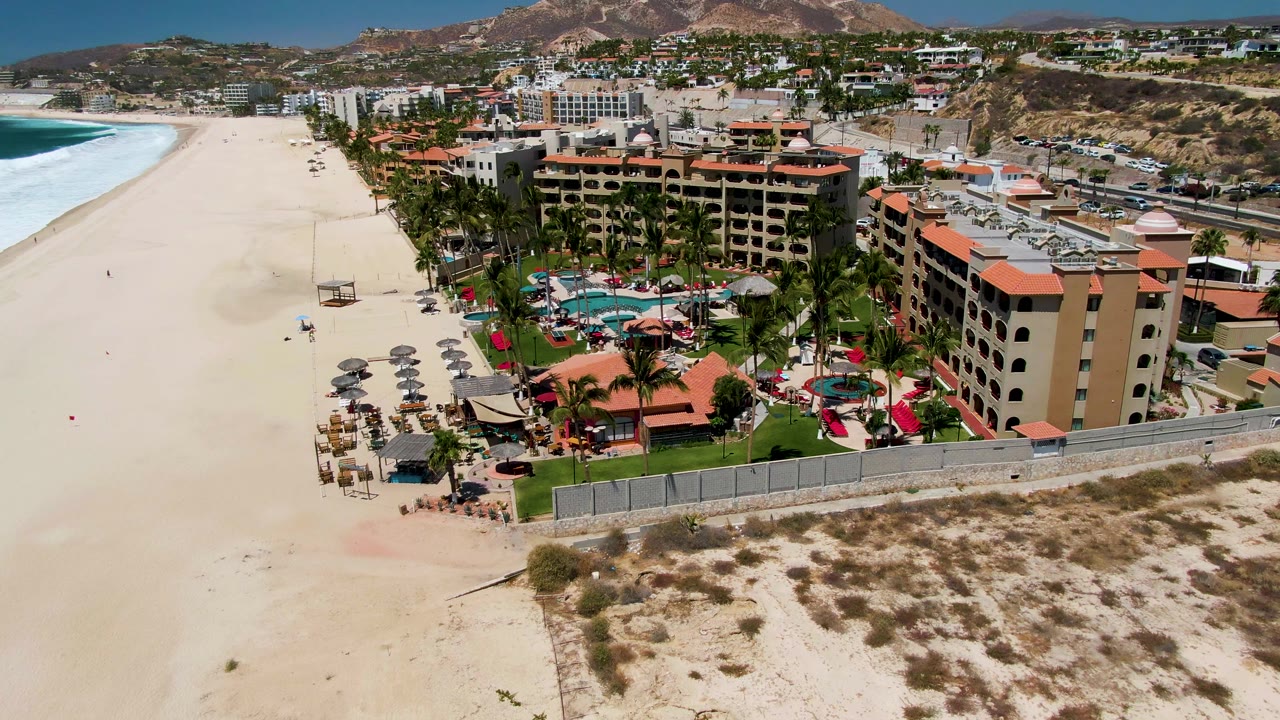 Aerial view in San Jose Del Cabo showing a wide sandy beach, blue surf on the left, resort buildings with red roofs, palms, pools, and small visible beachgoers near the shoreline.