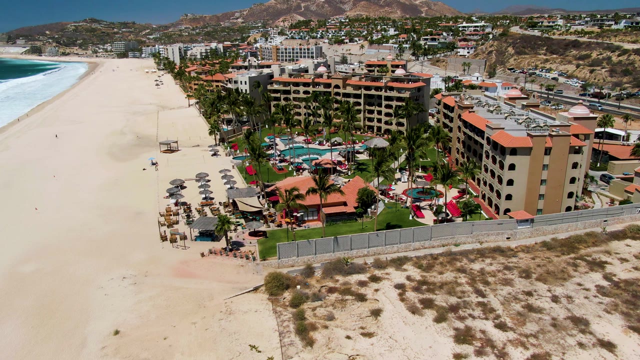 Wide beach aerial with resort pool area, palms, umbrellas, and ocean waves.