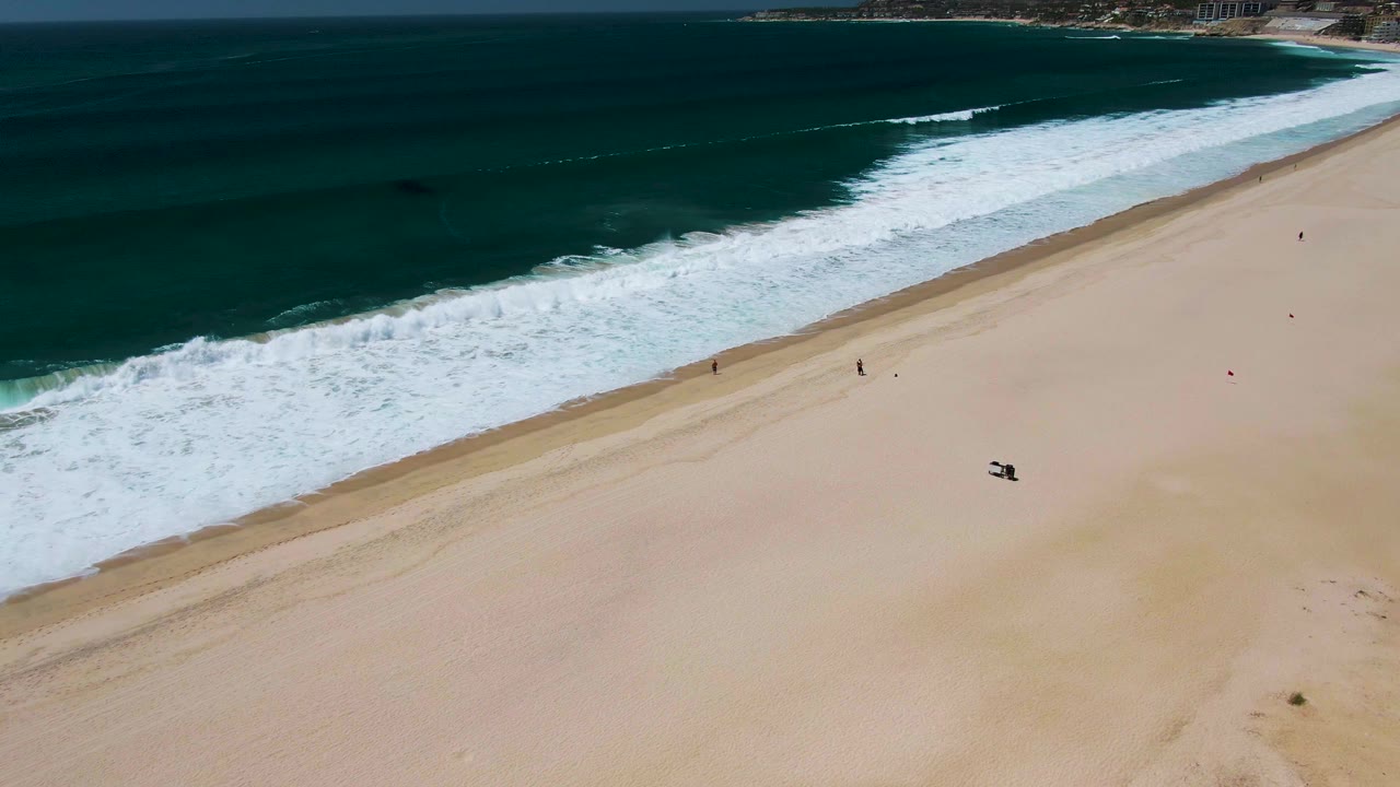 Aerial view over San Jose del Cabo surf with white waves crossing a wide sandy beach and tiny beachgoers visible near the shoreline.