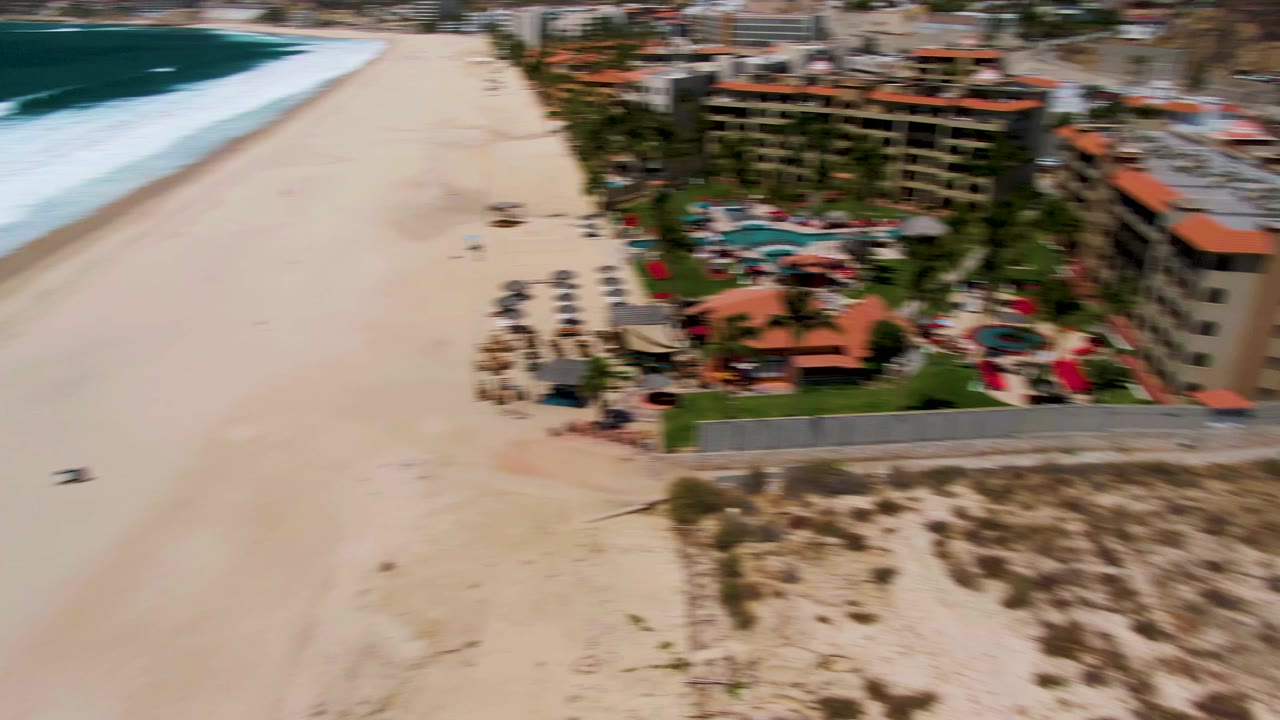 Aerial San Jose del Cabo beach view with resort buildings, pool areas, umbrellas, palm trees, dune scrub, and pale sand beside the surf.