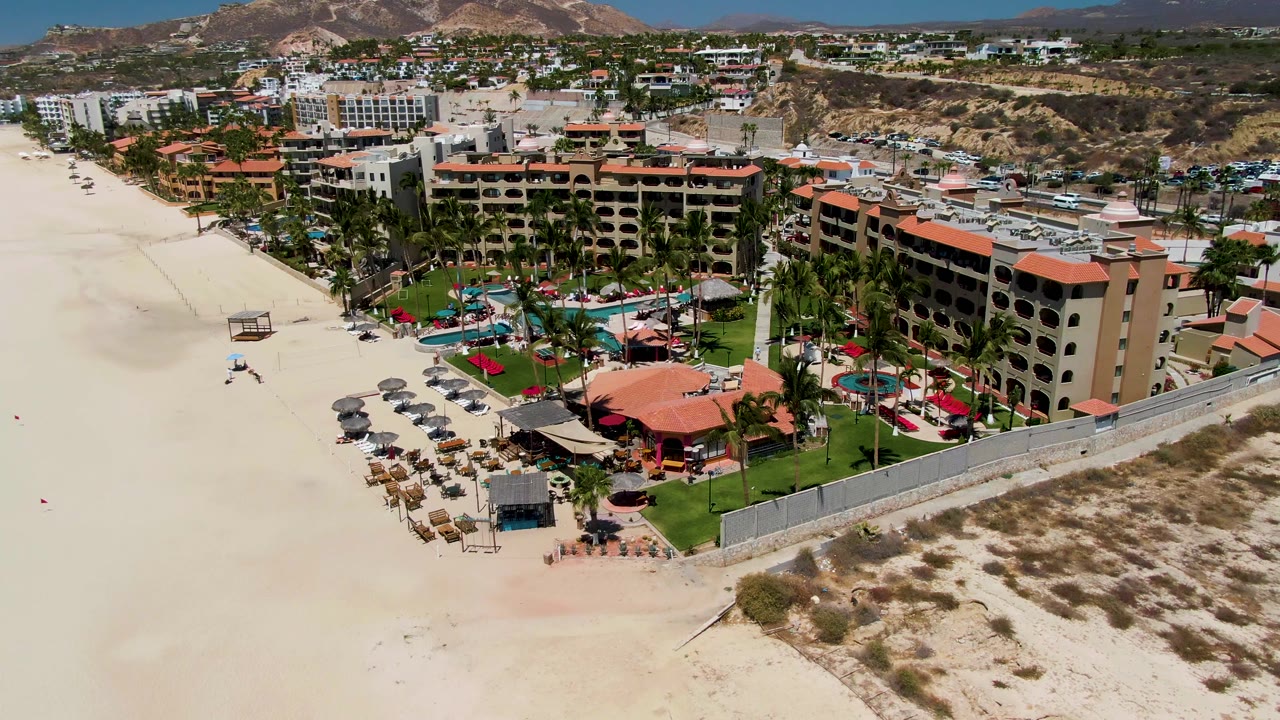 Aerial frame in San Jose Del Cabo showing a beachfront resort with red rooflines, pool area, palms, lounge chairs, wide sand, town buildings, and hills beyond.