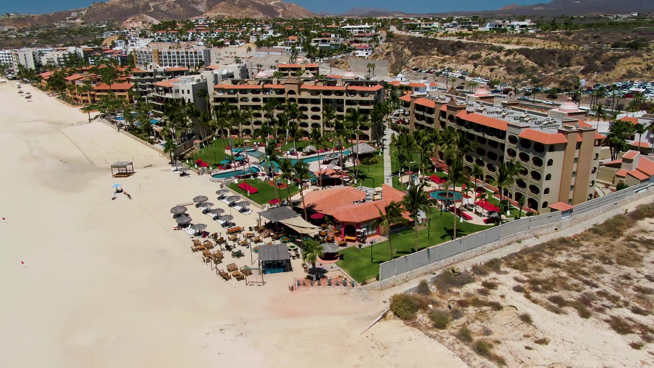 Drone view over sand, resort buildings, pool area, palms, and hills.