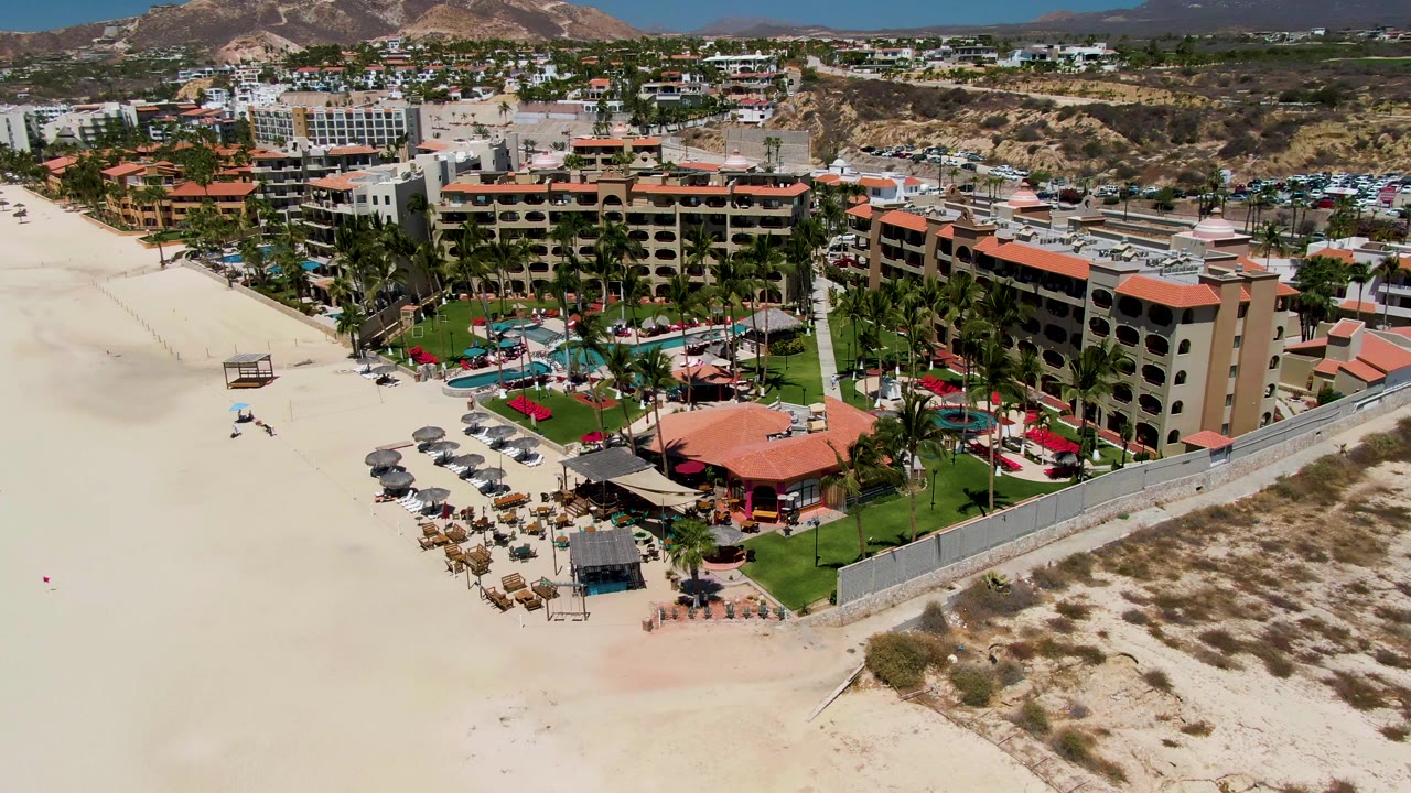 Aerial still from San Jose Del Cabo showing the resort closer to the right side of frame, with beach seating and umbrellas below, pools and palms inside the grounds, and mountains in the distance.