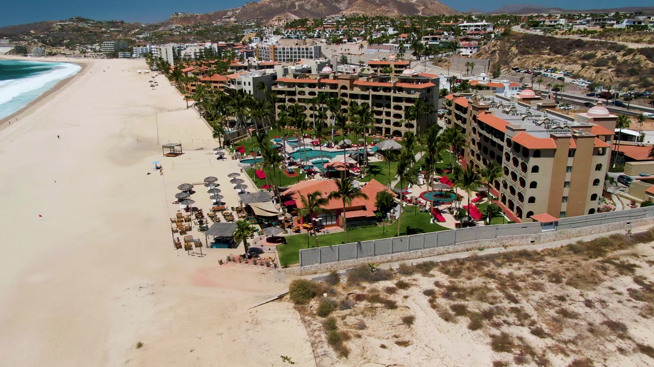 Aerial view in San Jose del Cabo showing a broad sandy beach, blue-green surf at left, a resort complex with red roofs at right, and small visible beachgoers near chairs and umbrellas.