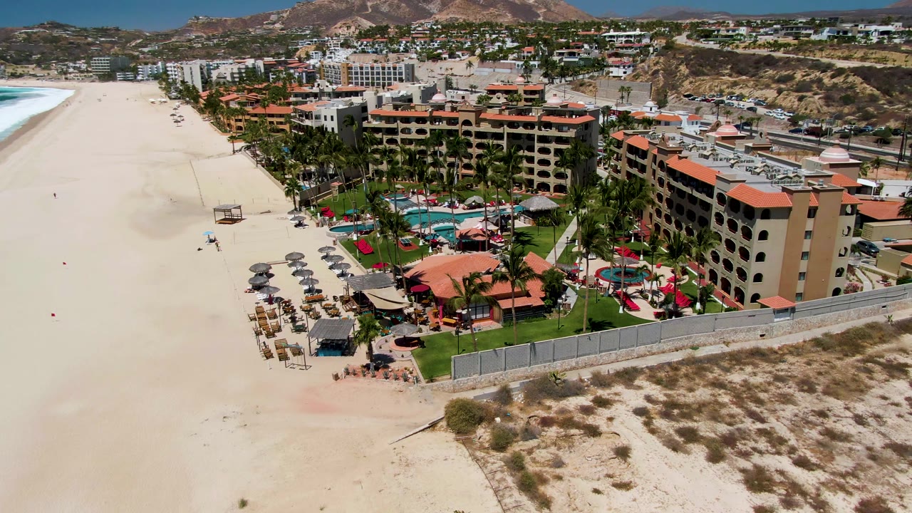 Aerial frame in San Jose del Cabo centered on the resort pool grounds, beach chairs and umbrellas on the sand, ocean surf at left, hillside homes and buildings in the background, and small visible people.