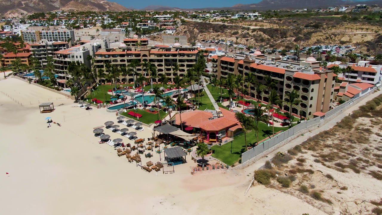 Aerial view in San Jose del Cabo, Mexico, showing a beachfront hotel complex beside pale sand, pool courtyards, umbrellas, palm trees, and hills in the distance.