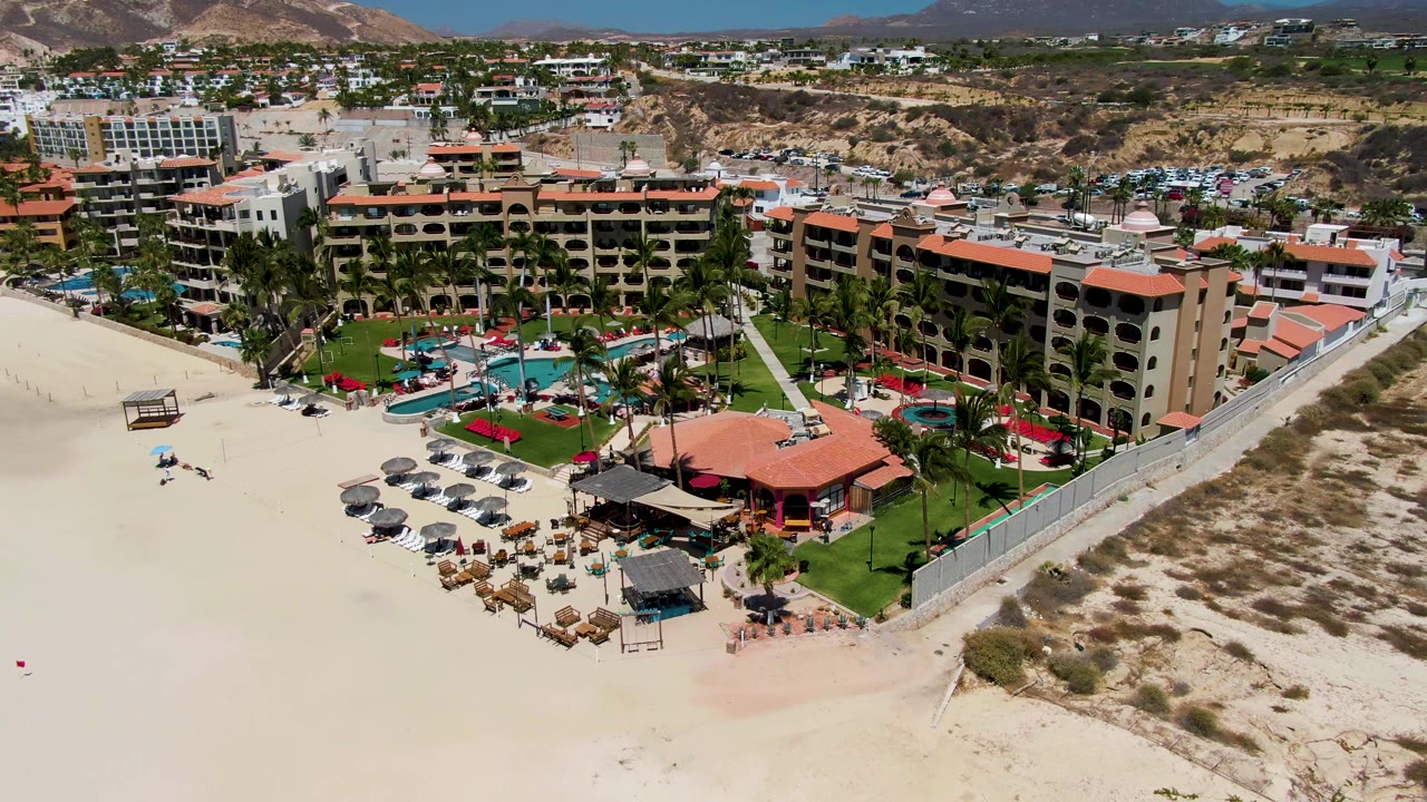 High aerial frame over San Jose del Cabo resort buildings with red tile roofs, green landscaping, turquoise pools, beach umbrellas, open sand, and low hills behind the town.