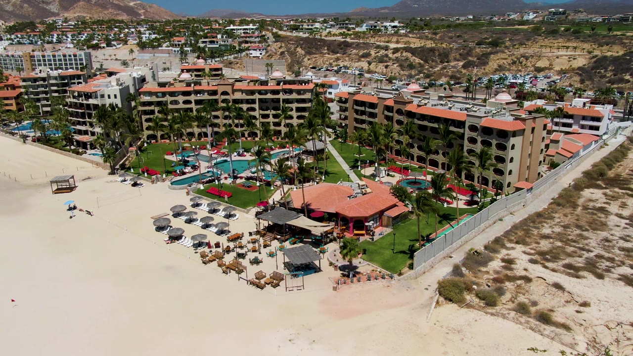 Drone-height view in San Jose del Cabo with the hotel complex, palm-lined pool area, rows of umbrellas on the beach, scrubby dunes, distant buildings, and hills under bright light.