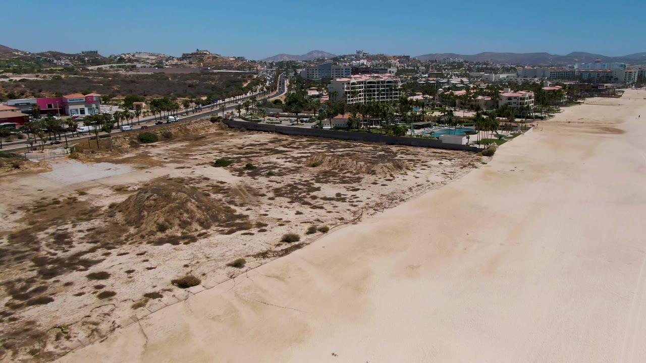 Opening aerial frame over San Jose del Cabo shows scrubby dunes in the foreground, wide pale beach to the right, a coastal road with cars, low buildings, palms, distant hills, and no visible people.