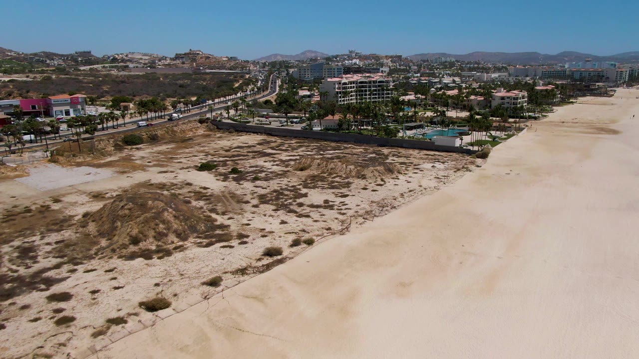 At one second, the San Jose del Cabo aerial holds the dune field lower in frame, the beach runs along the right, cars move on the road, buildings and palm trees sit beyond, and no visible people appear.