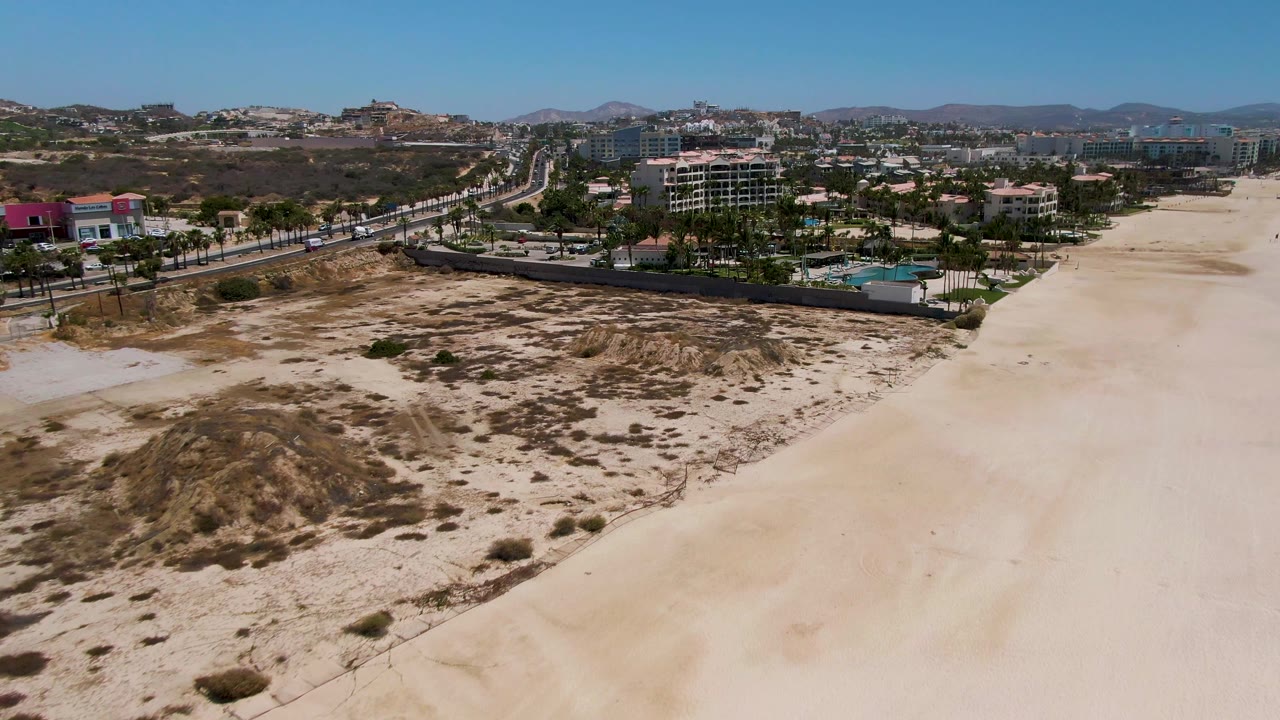 At two seconds, the aerial view over San Jose del Cabo shows more of the broad sand strip on the right, scrub patches across the dune area, roadside cars, low-rise buildings, palms, hills, and no visible people.