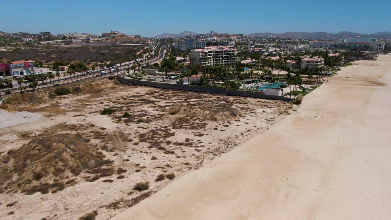 Aerial coastal landscape with dunes, beach, roadway, cars, buildings, palms, and hills.