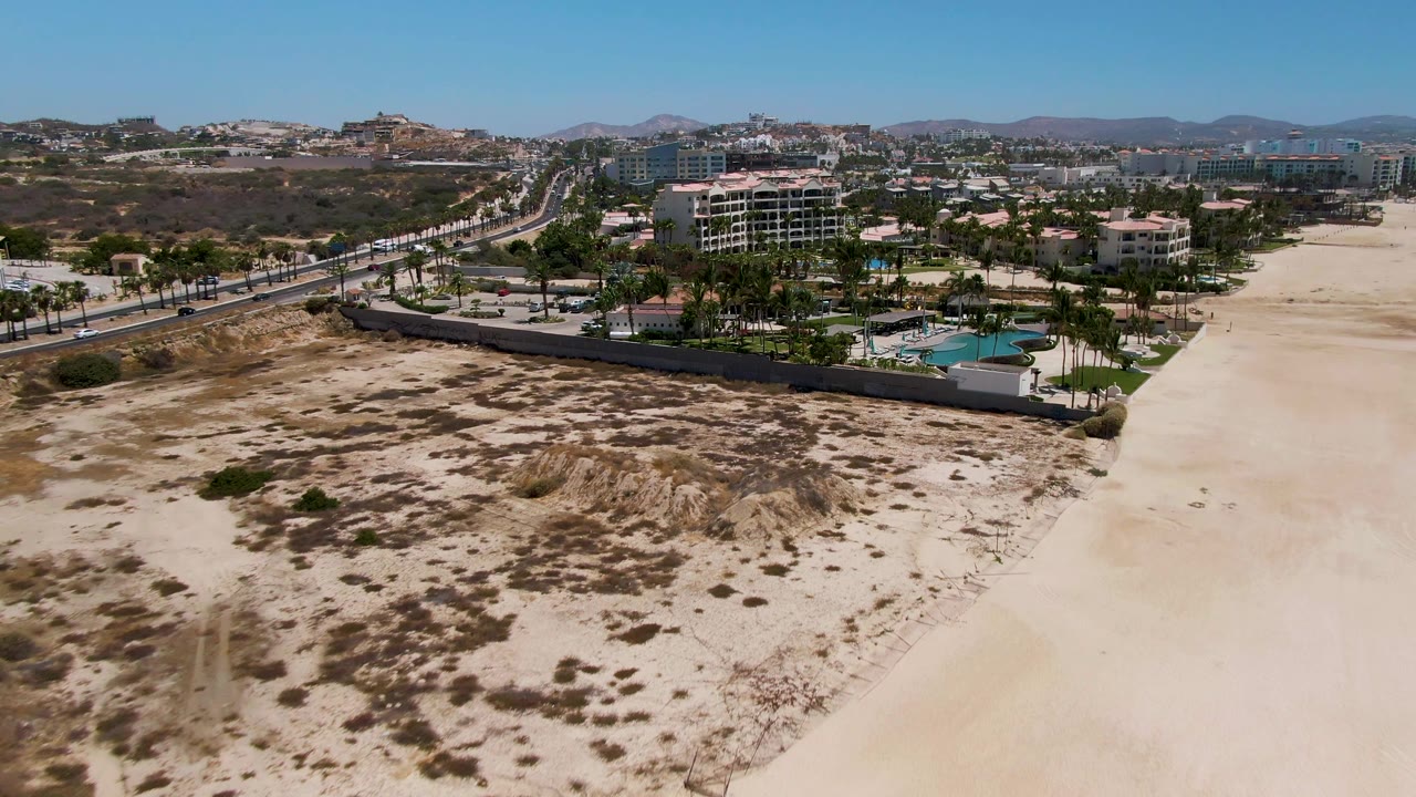 Opening aerial frame in San Jose del Cabo, Mexico, showing pale sand dunes in front of a walled resort with palms, a blue pool, a road with vehicles, distant buildings, hills, and no visible people.