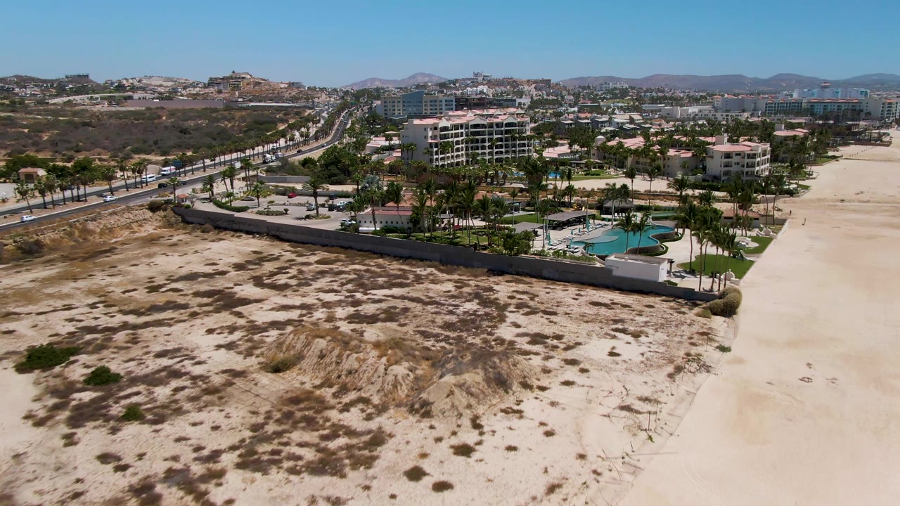 San Jose del Cabo aerial frame showing a broad stretch of pale beach sand and scrubby dunes beside a resort complex, with a curved pool, palms, road traffic, distant buildings, and no visible people.