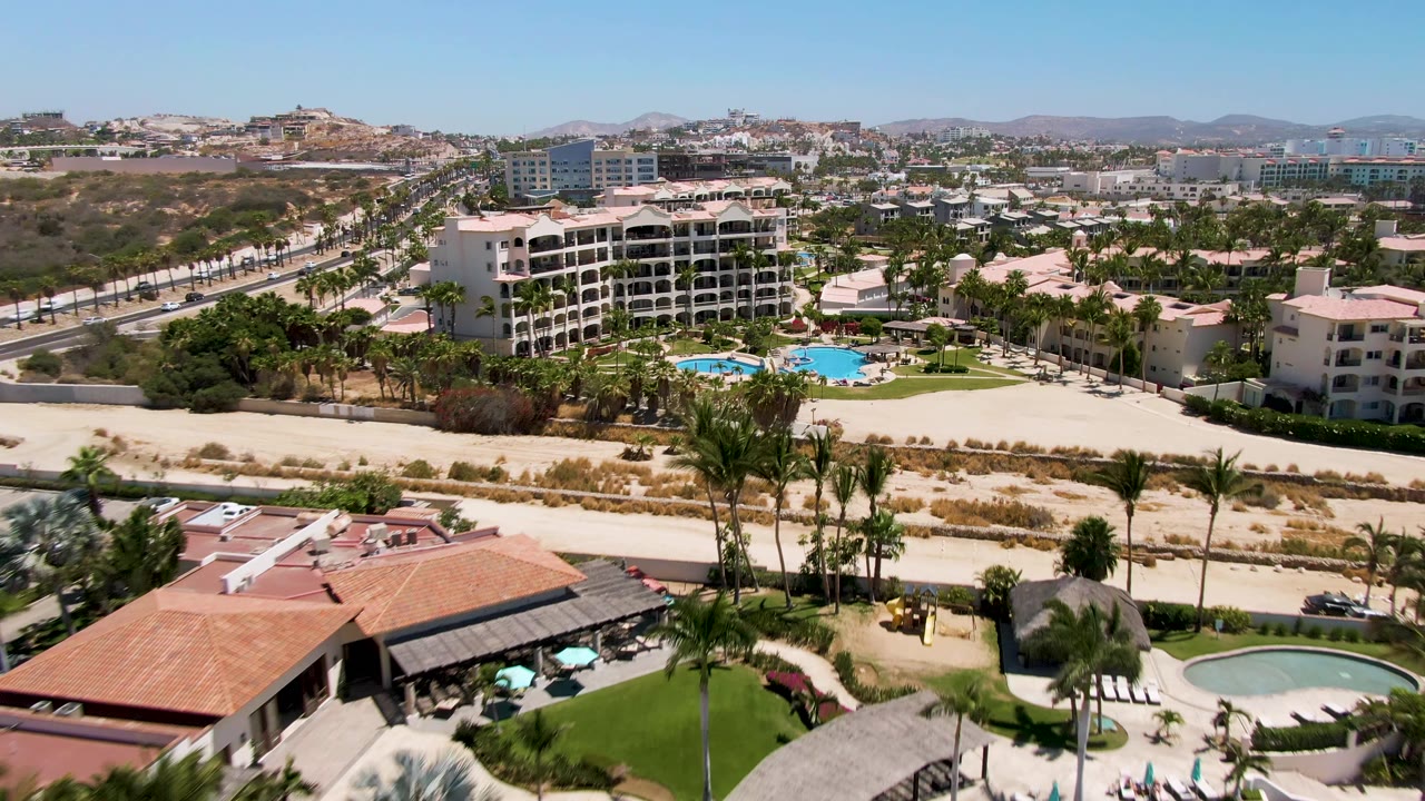 Aerial frame over San Jose del Cabo resort grounds with turquoise pools, palm trees, a parallel road, pale sand, low-rise buildings, distant hills, and no visible people.