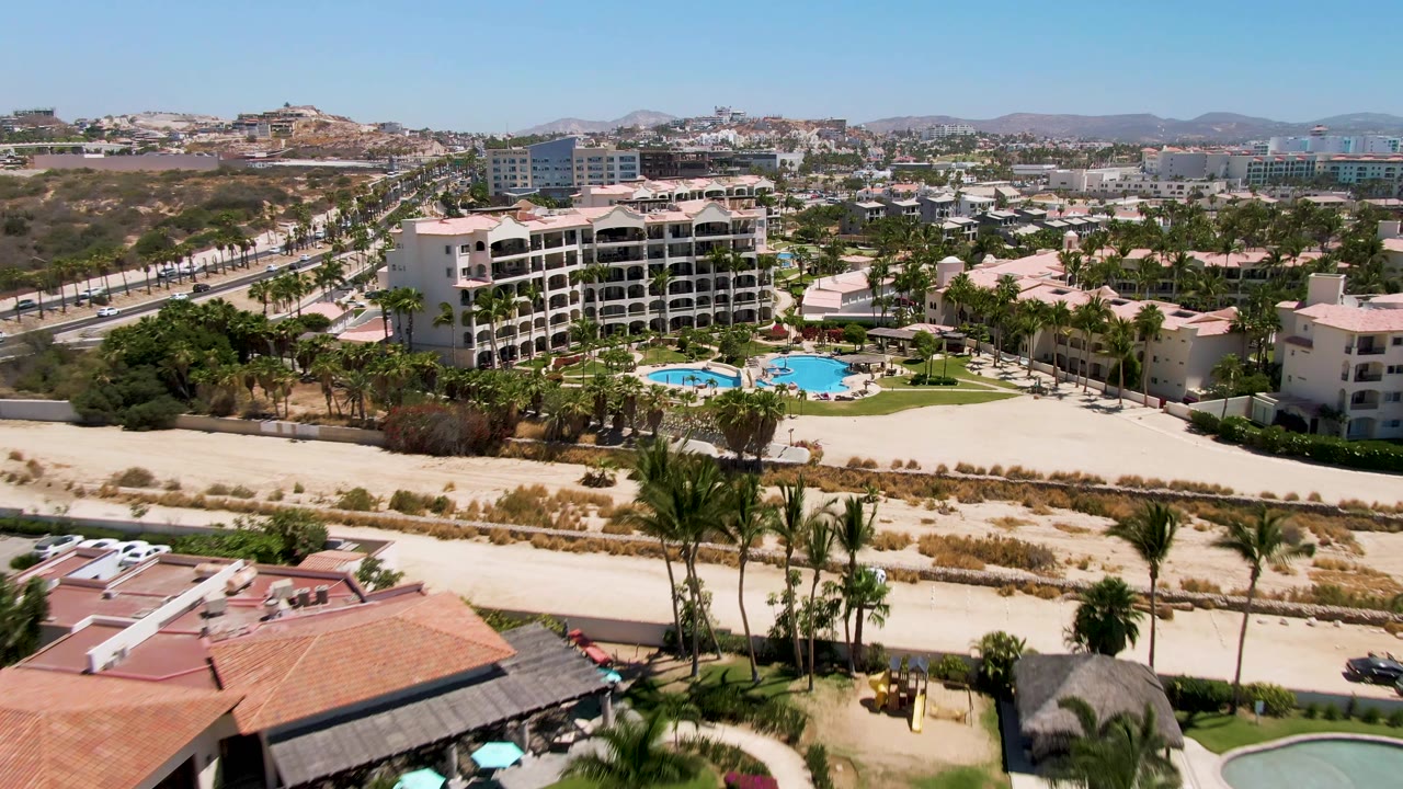 Aerial frame in San Jose del Cabo showing a palm-lined resort complex, bright blue pool water, sandy foreground, road lanes, distant buildings, hills, and no visible people.