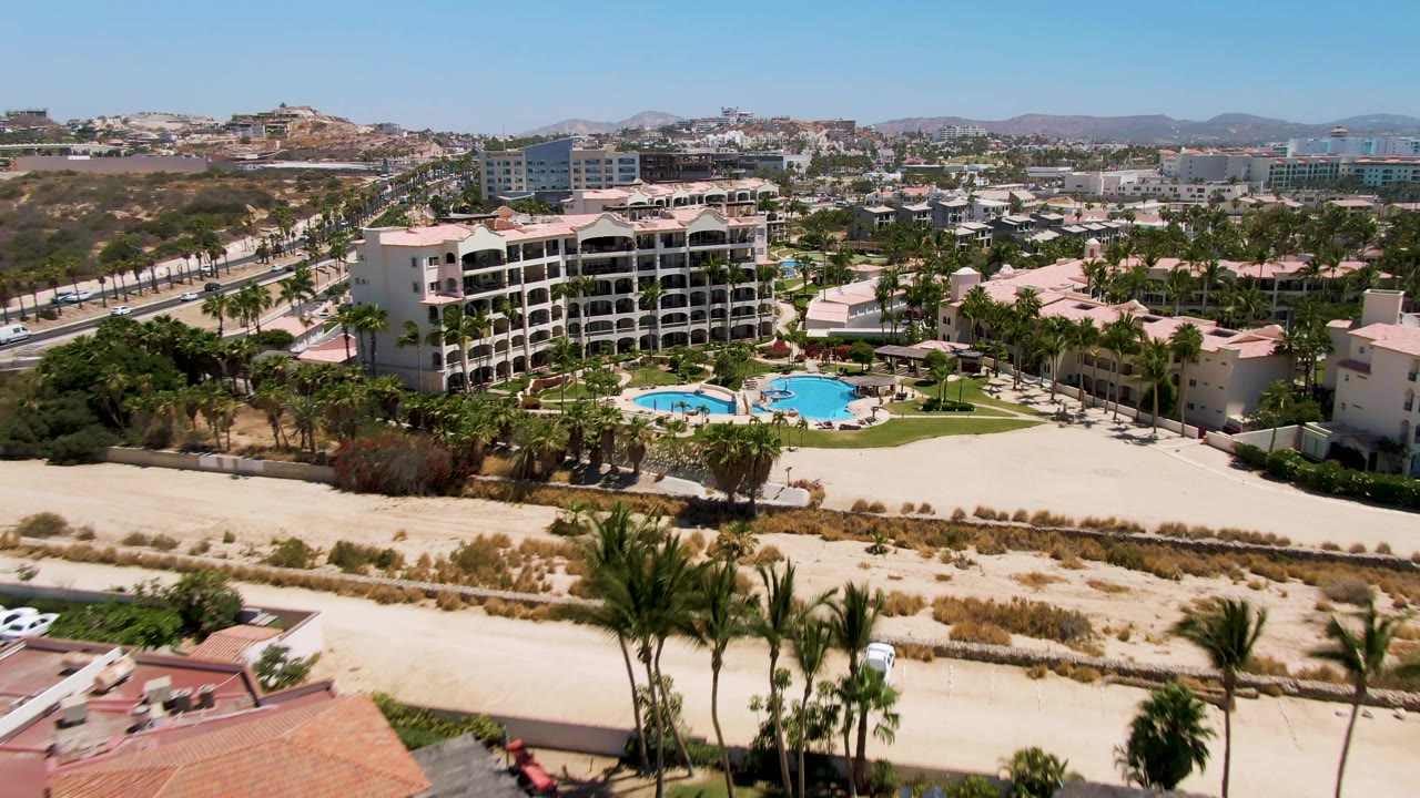 Aerial frame over San Jose del Cabo with resort pools and palms in the midground, sandy ground and road below, city buildings beyond, distant hills, and no visible people.