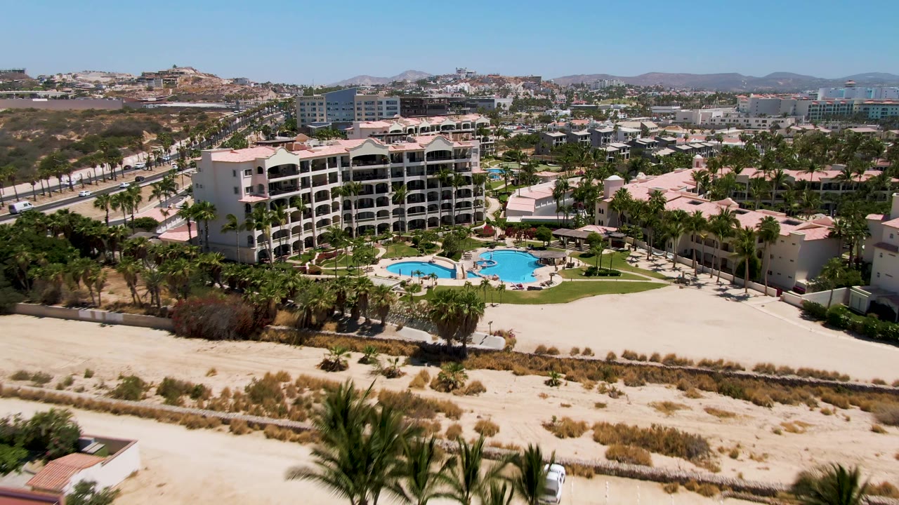 Aerial resort scene with pools, palms, road, sand, and buildings under clear sky