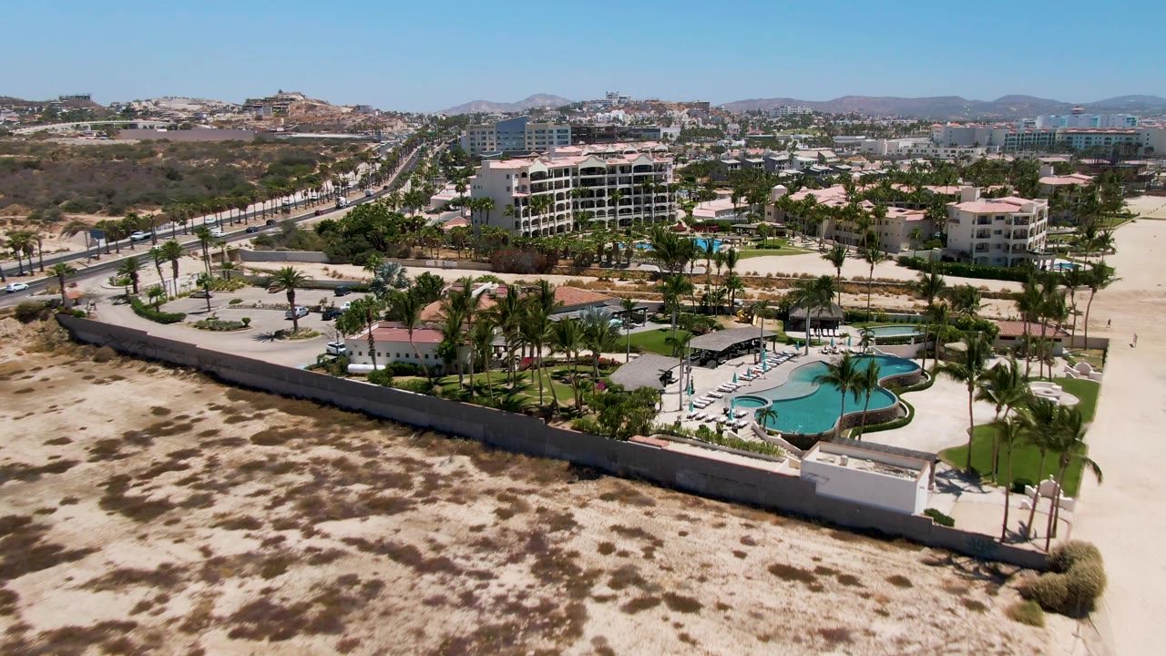Wide aerial frame over San Jose Del Cabo showing sandy foreground, a resort pool area with palms, a road at left, distant buildings, and no visible people.