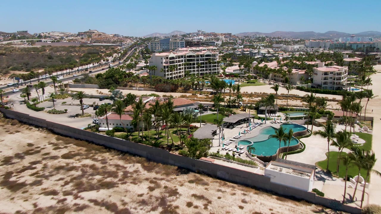San Jose Del Cabo aerial view with the curved turquoise pool closer to center, palm landscaping below, sandy open ground, road traffic at left, and no visible people.
