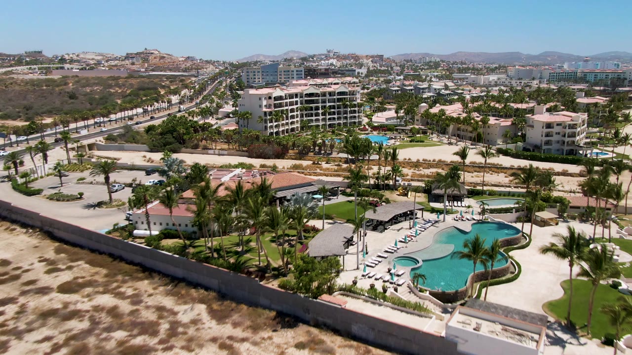 Aerial frame in San Jose Del Cabo showing the resort pool and palms at lower right, sandy terrain across the foreground, roads and buildings behind, and no visible people.