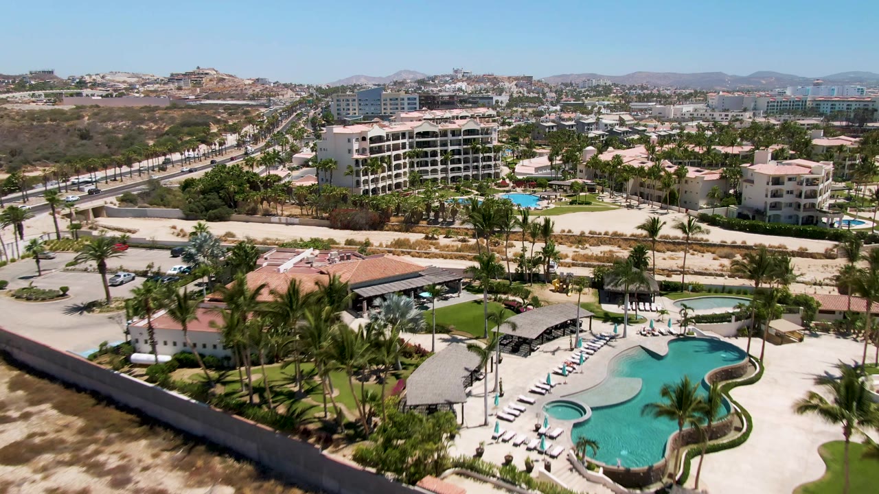 Closer aerial view of resort pool, loungers, palms, road, sandy strip, and hotel buildings.