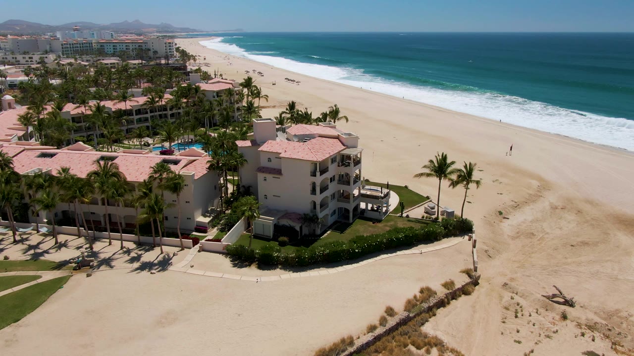 Aerial view of resort buildings, palm trees, sandy beach, and ocean surf.