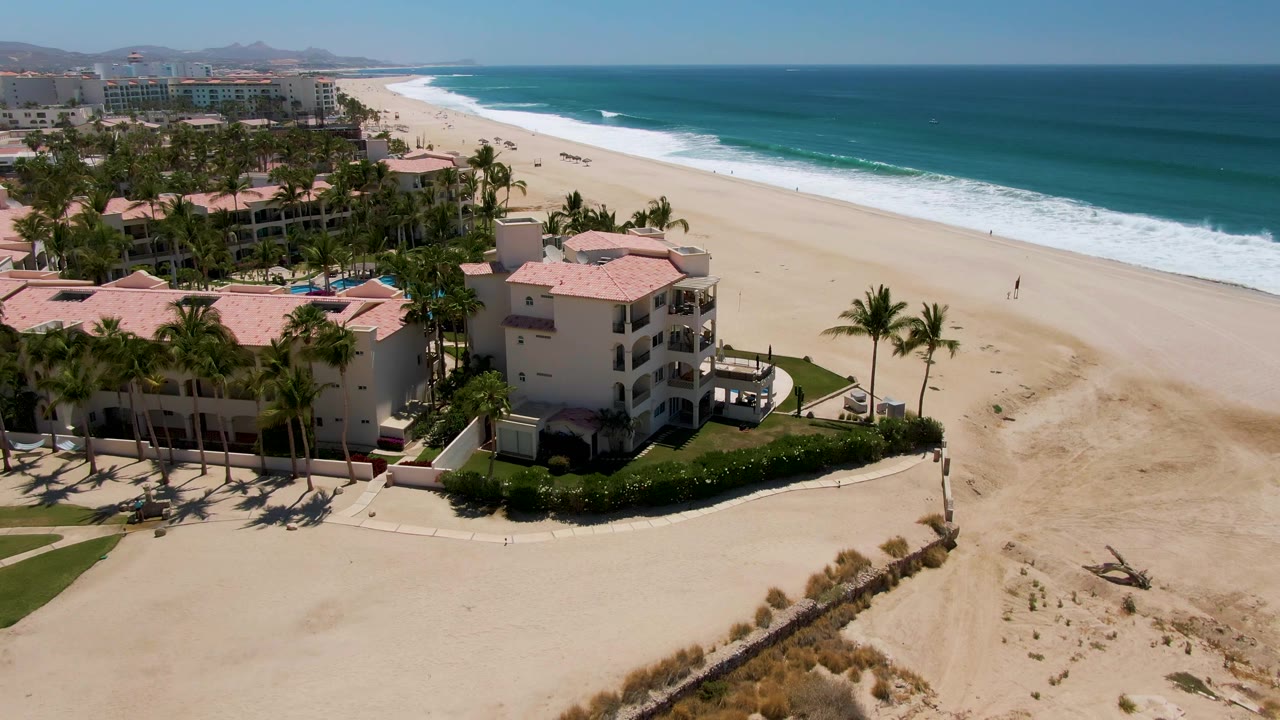 Aerial frame over San Jose del Cabo showing the resort shifted slightly in view, with palm shadows, a beach path, open sand, and white waves breaking near shore.