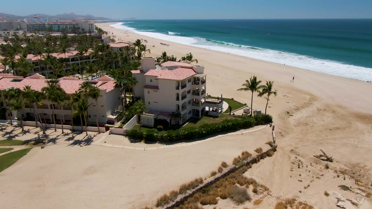 San Jose del Cabo aerial frame with the resort closer to center, a lone palm near the sand, low dunes in the foreground, and parallel surf lines on the blue water.
