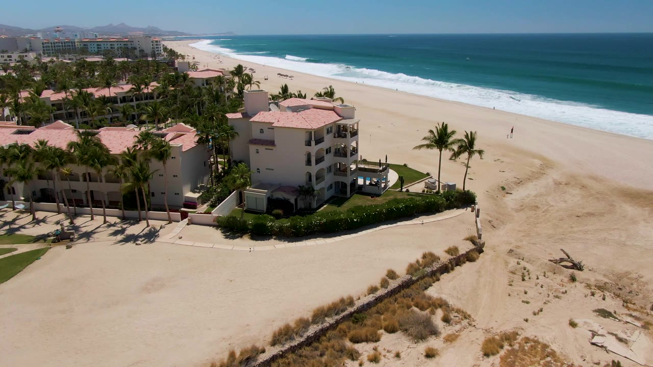 Later aerial frame over San Jose del Cabo with palm-lined resort buildings filling more of the left side, dune grass in the foreground, and the ocean horizon running across the right side.