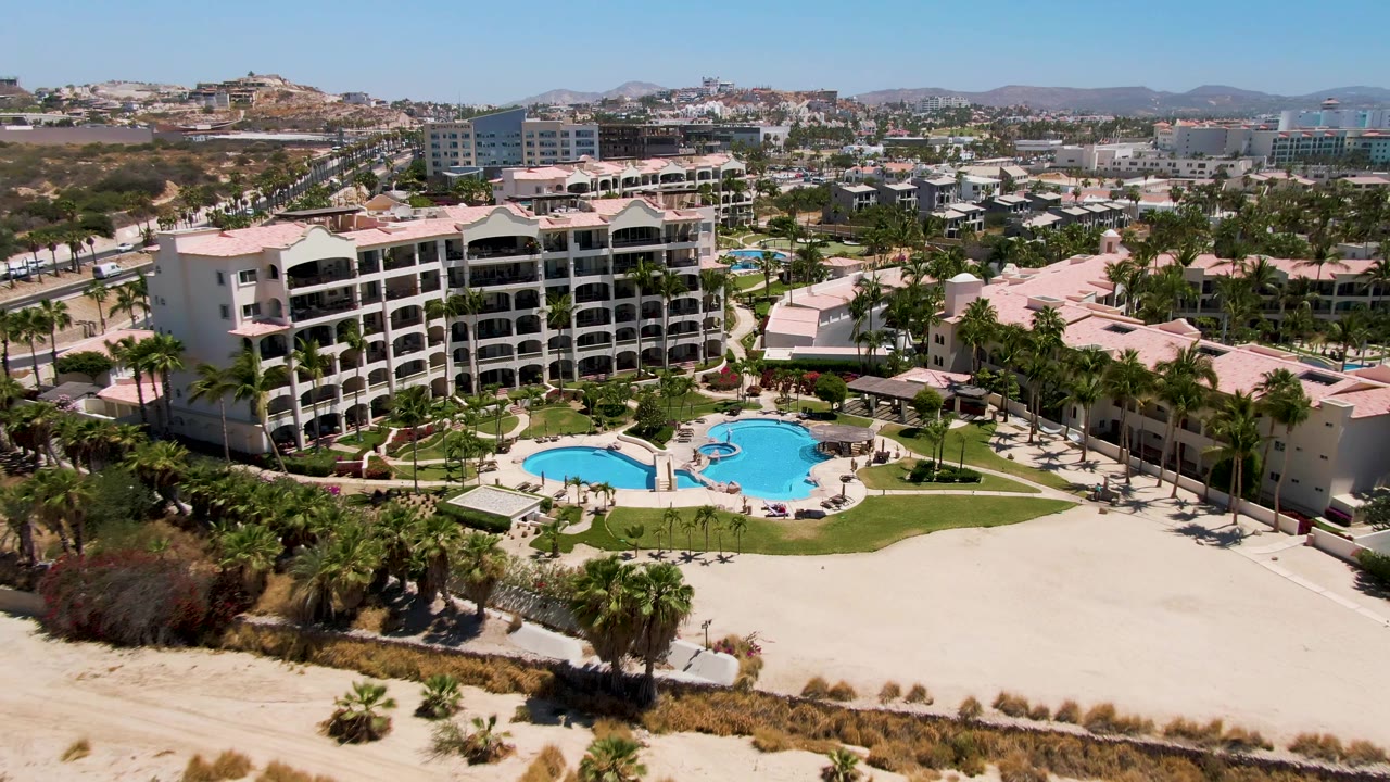 Aerial opening view over a San Jose Del Cabo resort with a blue pool, pale sand foreground, white buildings, palms, distant town, and no visible people.