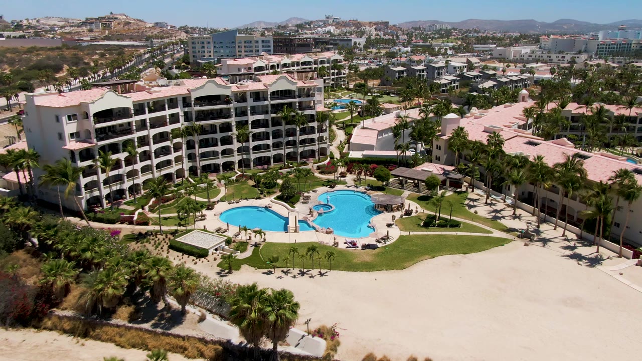 Aerial frame over San Jose Del Cabo with the white resort buildings and blue pool filling more of the center, sand and palms below, and no visible people.