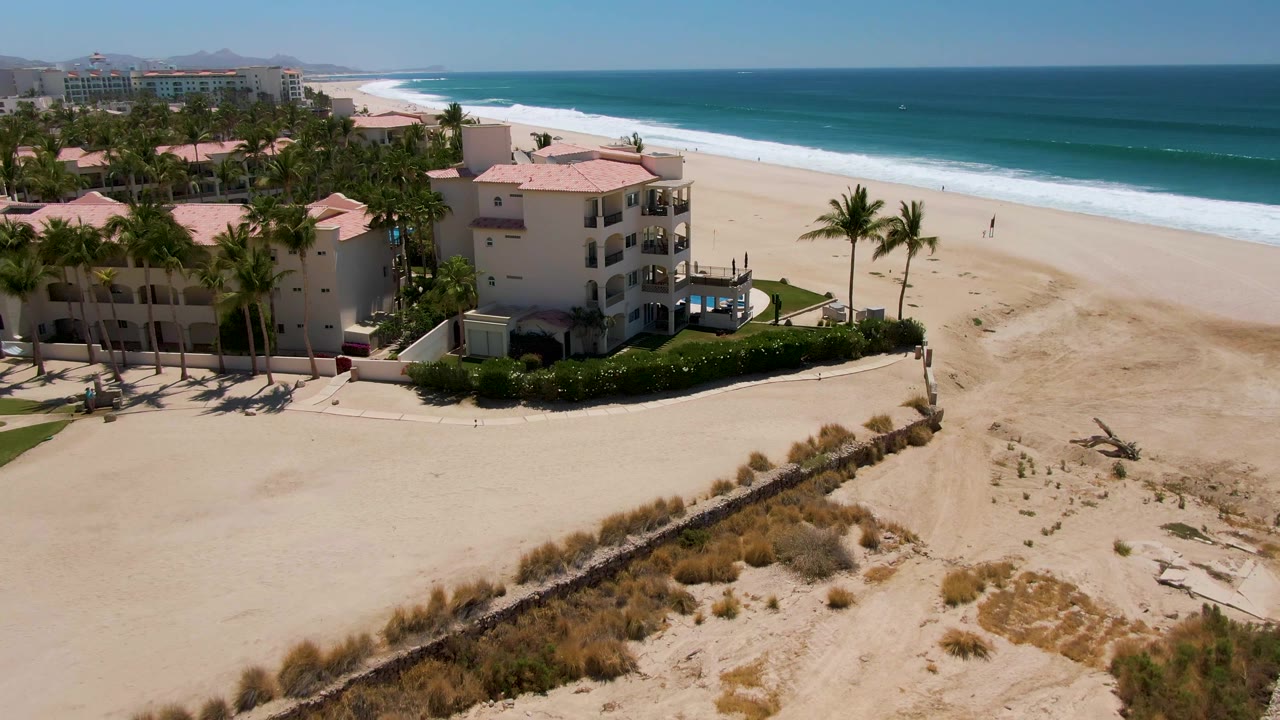 Aerial frame over San Jose Del Cabo beachfront with hotel buildings and palm trees at left, dunes in front, blue ocean beyond the wide sand, and small people visible near the surf.