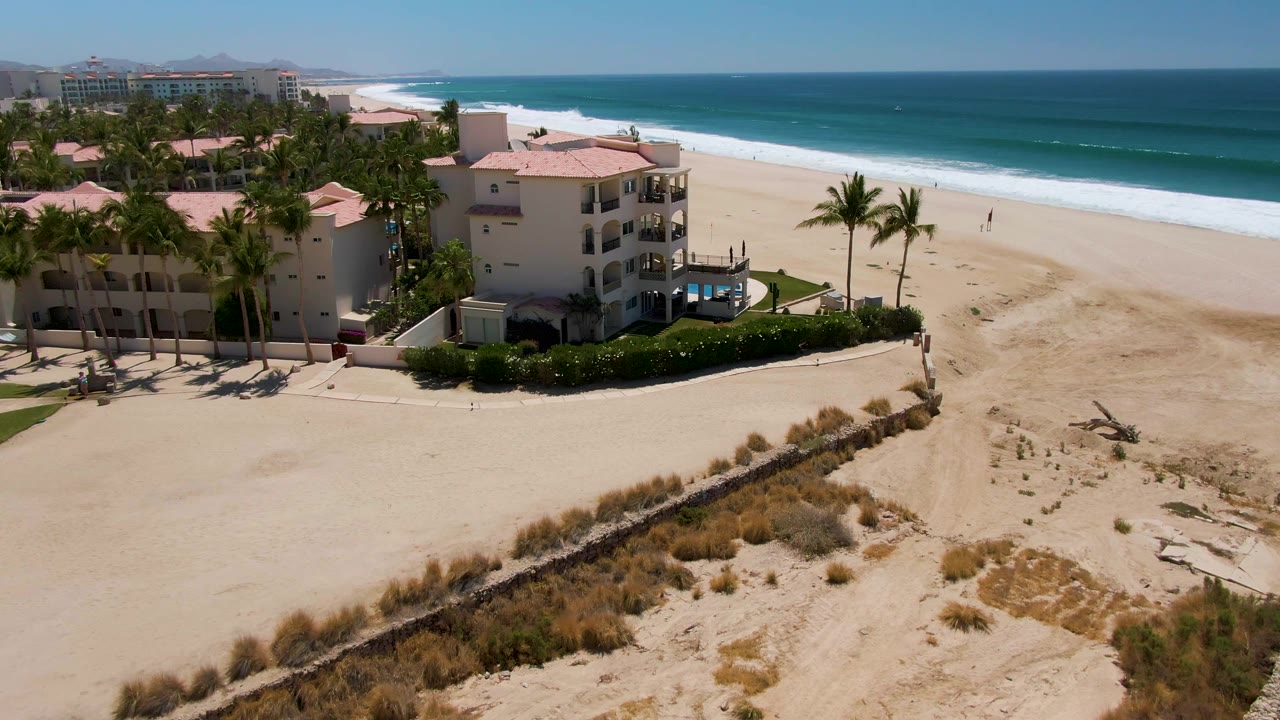 Beachfront resort buildings and palms beside a wide sandy beach and blue surf.