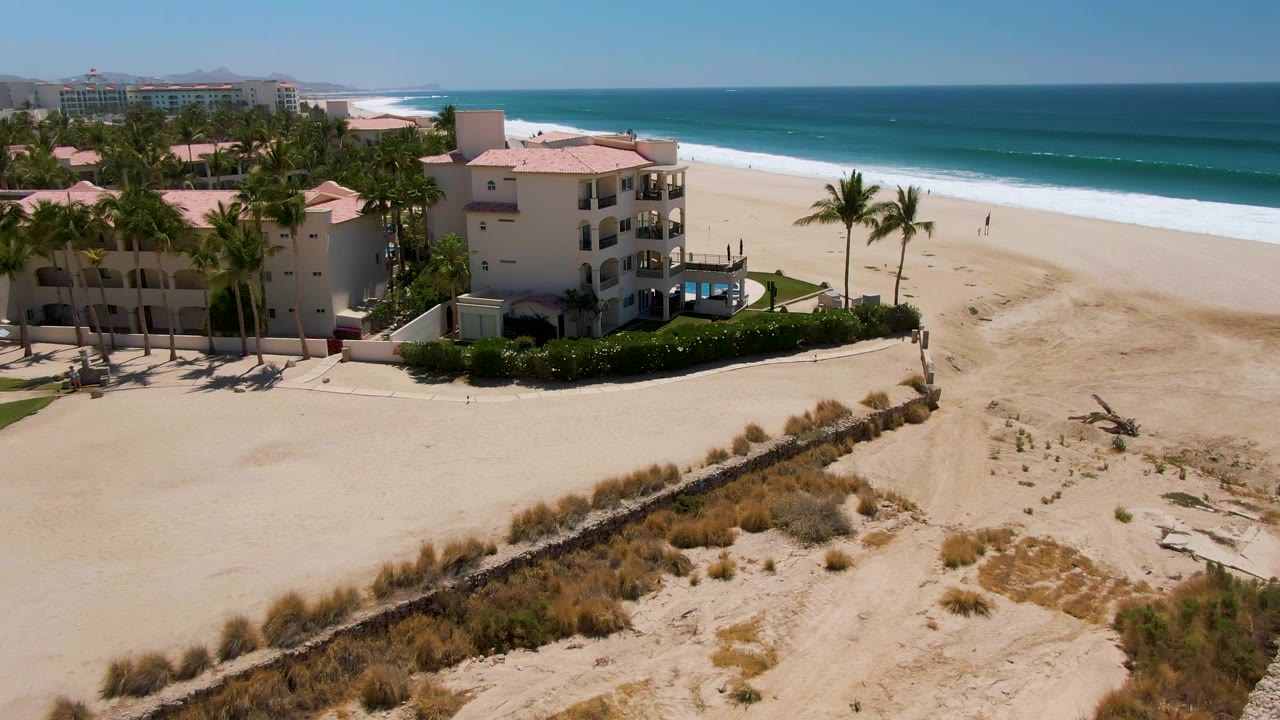 San Jose Del Cabo aerial frame with the hotel complex centered left, a curved wall and dune vegetation in the foreground, a mostly open beach, and blue ocean with a thin white surf band.
