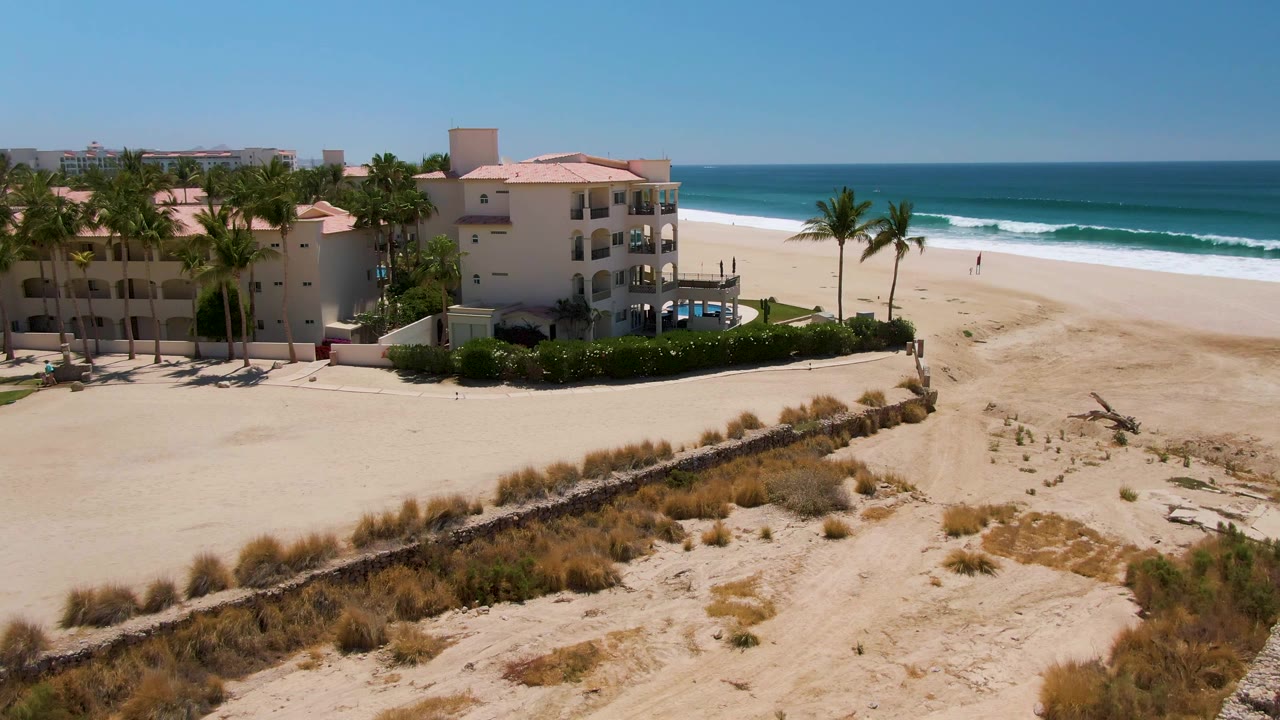Elevated view in San Jose del Cabo showing pale dunes in front, a cream beachfront building with palm trees on the left, and blue ocean waves breaking on the sandy beach to the right.