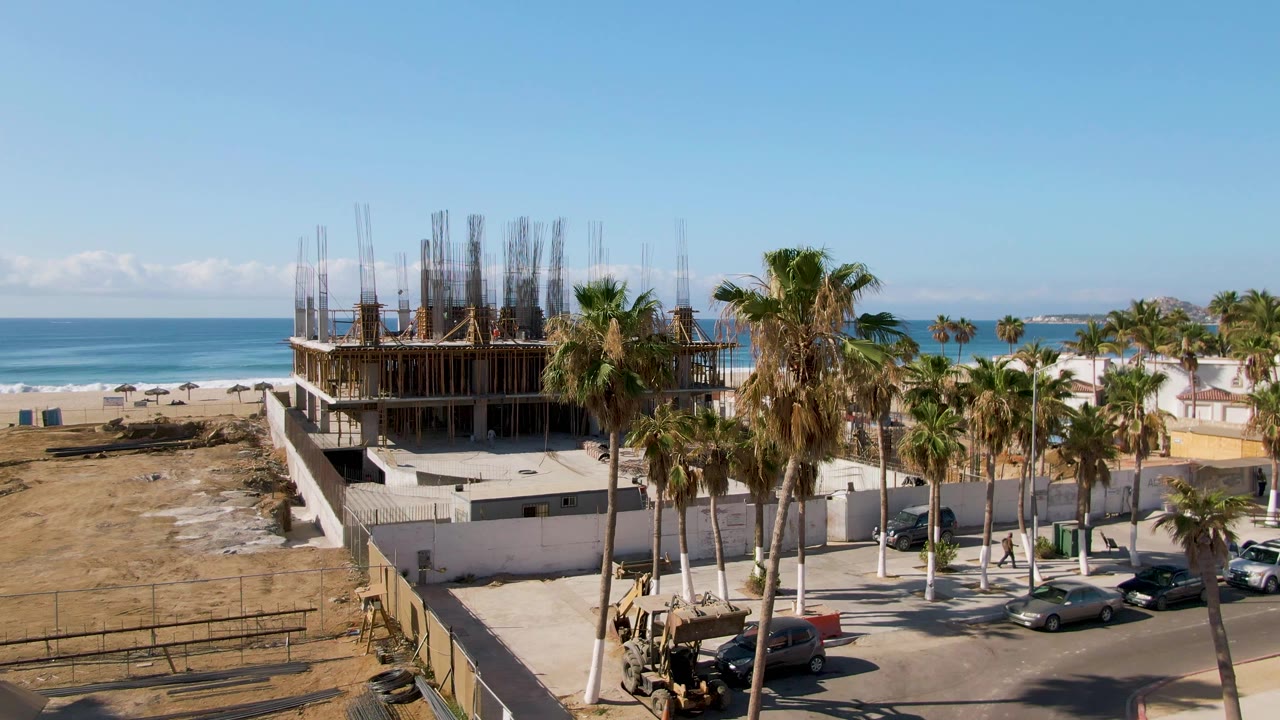 Wide aerial view at El Tule Beach Los Cabos showing an unfinished concrete building with rebar, palms, parked cars, sand, blue ocean, and no visible people.