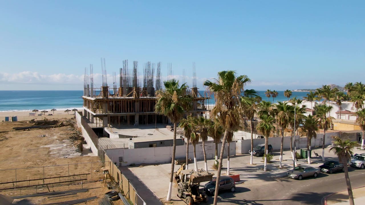 Aerial frame at El Tule Beach Los Cabos with the unfinished building centered, a white perimeter wall below it, palms to the right, ocean in back, and no visible people.