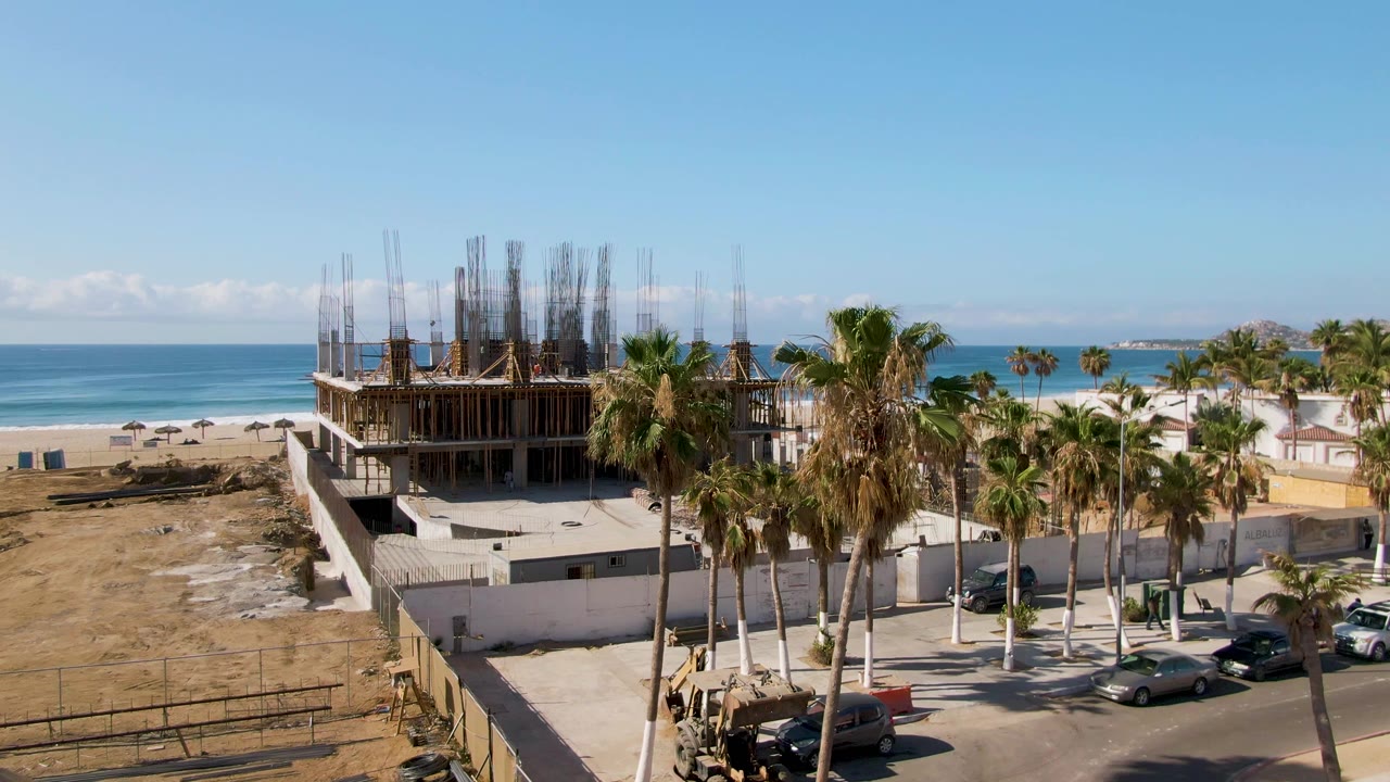 El Tule Beach Los Cabos aerial view showing the building under construction, a cluster of palms, several parked cars, sandy ground, ocean waves, and no visible people.